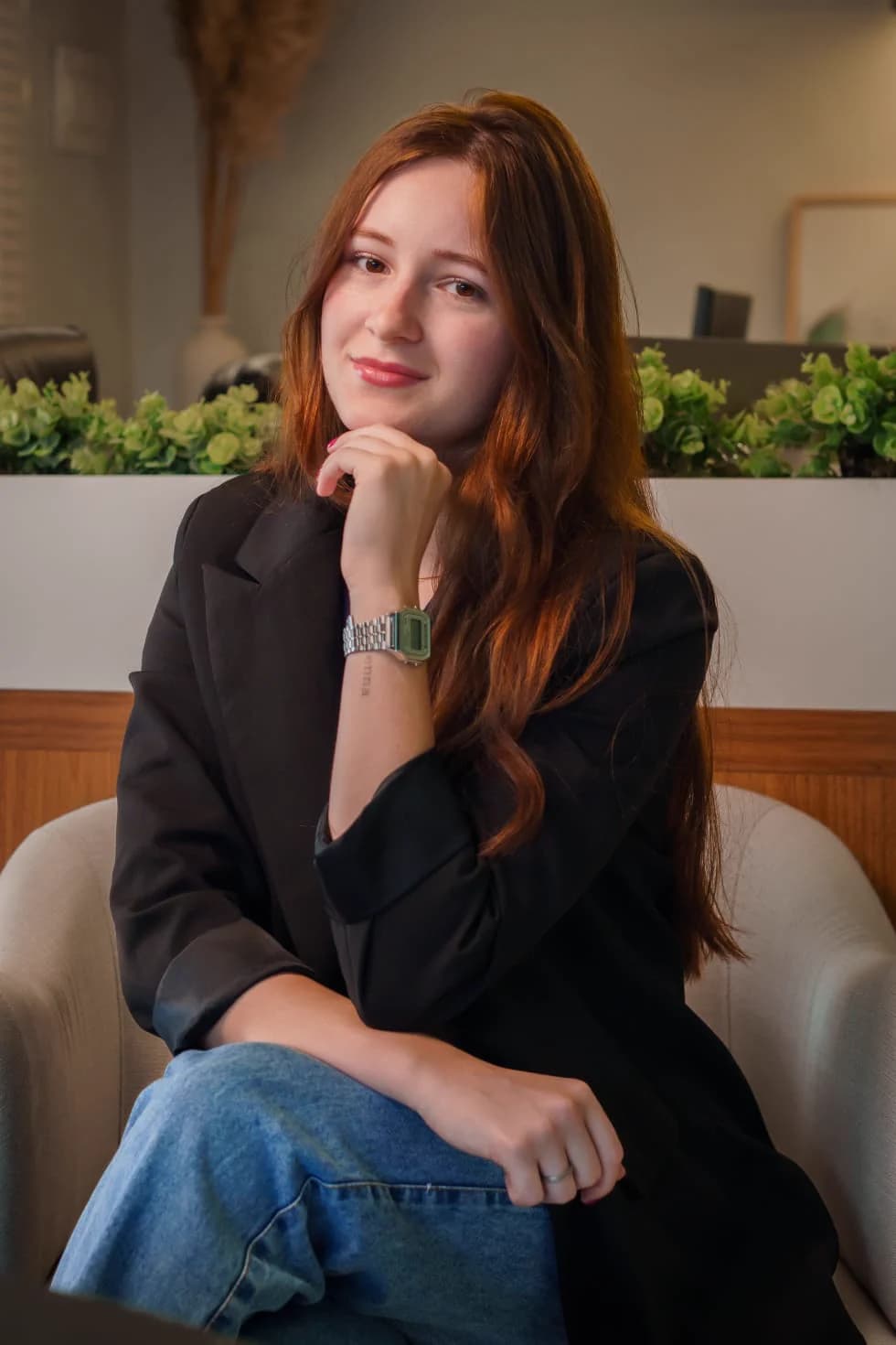 Adriana sentada no escritório, de blazer preto, em um ambiente com plantas.